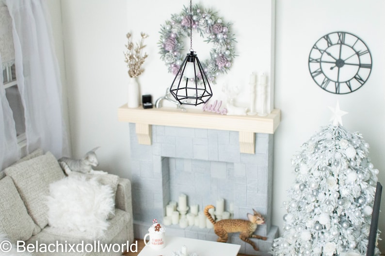 May include: A miniature dollhouse living room with a white fireplace, a white Christmas tree decorated with silver ornaments, a white couch, and a black pendant light hanging above a white wreath.