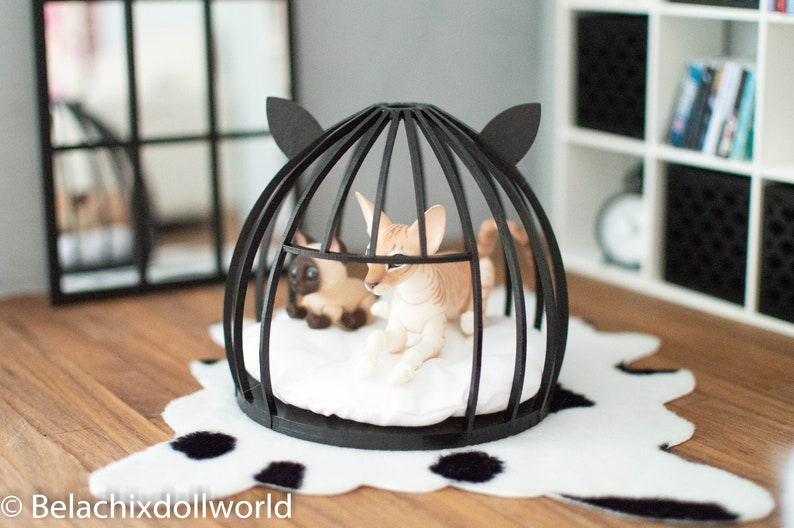May include: A black cat bed with a white cushion inside. Two small, white and brown cats are inside the bed. The bed is on a white and black cowhide rug.
