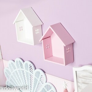 May include: Two white and pink wooden house-shaped shelves mounted on a light purple wall. The shelves have small cut-out windows and are perfect for displaying small items.