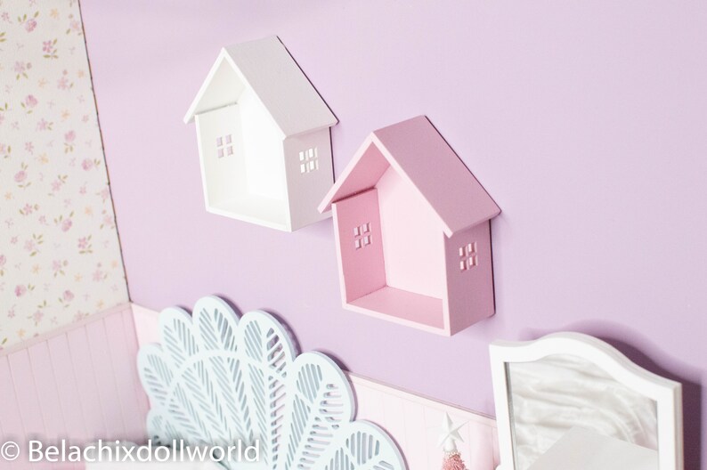 May include: Two white and pink wooden house-shaped shelves mounted on a light purple wall. The shelves have small cut-out windows and are perfect for displaying small items.