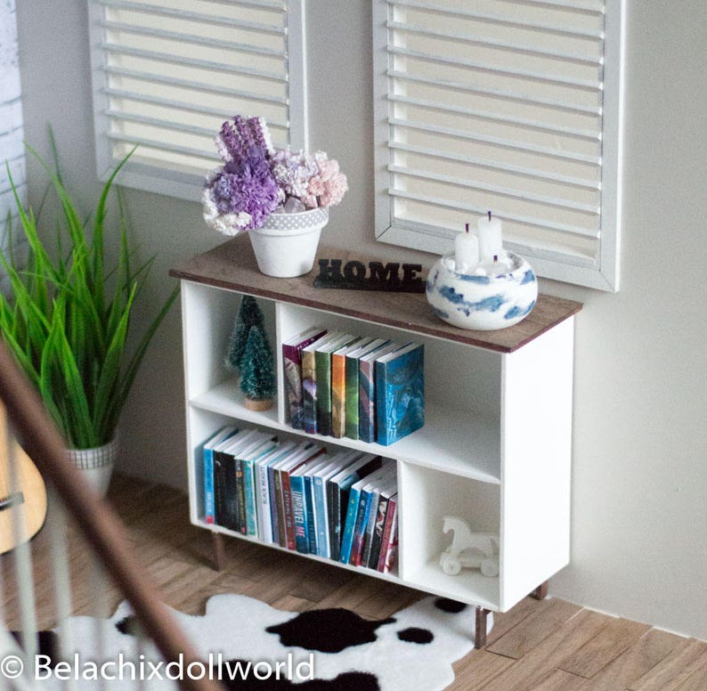 May include: White bookshelf with four shelves, filled with books and decorative items. The bookshelf has a wooden top with the word "HOME" in black letters. There is a potted plant with purple flowers on the top shelf, and a white ceramic planter with candles on the top shelf. There is a small artificial Christmas tree on the second shelf.