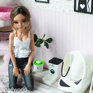 May include: A doll wearing a white tank top and blue jeans is kneeling in front of a white automatic self-cleaning litter box for cats. The litter box has a black base and a white lid. There is a green automatic pet water fountain and a black automatic pet food dispenser in the background.