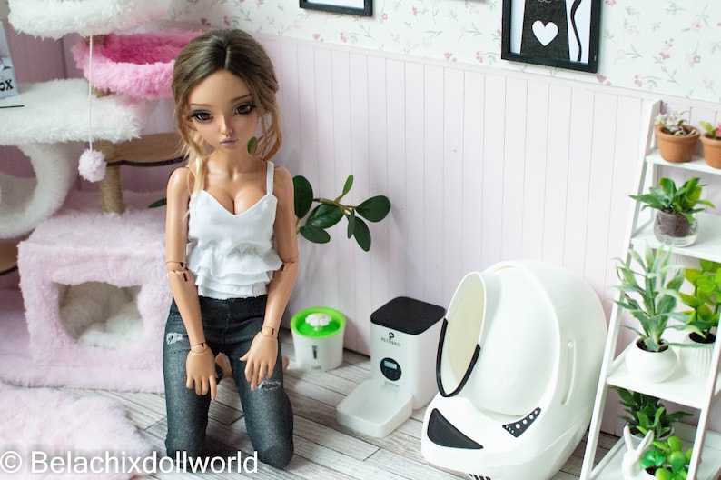 May include: A doll wearing a white tank top and blue jeans is kneeling in front of a white automatic self-cleaning litter box for cats. The litter box has a black base and a white lid. There is a green automatic pet water fountain and a black automatic pet food dispenser in the background.