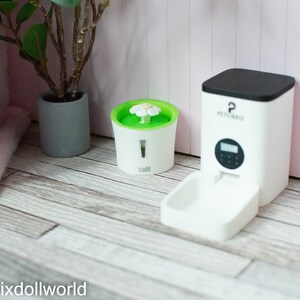 May include: White automatic pet feeder with a black top and a digital display. The feeder has a white bowl attached to the front. A white water fountain with a green top and a white flower on top is also shown.