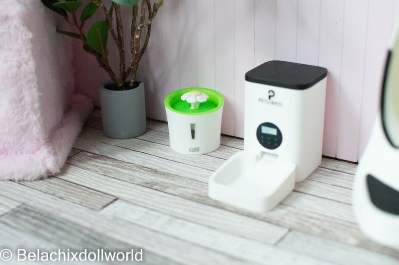 May include: White automatic pet feeder with a black top and a digital display. The feeder has a white bowl attached to the front. A white water fountain with a green top and a white flower on top is also shown.