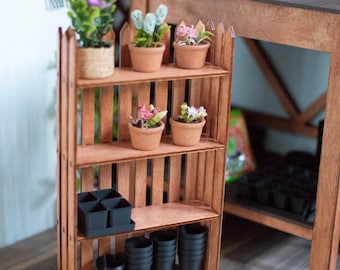 Puede incluir: Un estante de plantas de madera en miniatura con tres estantes, que contiene pequeñas plantas en macetas con flores moradas, rosas y azules. El estante también contiene macetas y una bandeja negras. La escena evoca un jardín en miniatura.