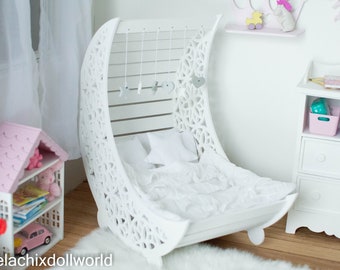 Op de afbeelding: Witte halvemaanvormig bed met een wit hoofdeinde en witte beddengoed. Het hoofdeinde heeft een decoratief uitgesneden ontwerp en een hangende mobiel met sterren en harten.