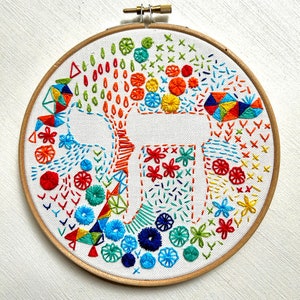 Puede incluir: Un bastidor de bordado colorido con un lienzo blanco que presenta una gran letra estilizada y varios diseños bordados. El bordado incluye formas geométricas, flores y patrones abstractos en tonos de rojo, naranja, amarillo, verde y azul.