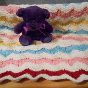 May include: A knitted baby blanket with a wavy pattern in white, pink, yellow, blue, and red. A purple teddy bear sits on top of the blanket.