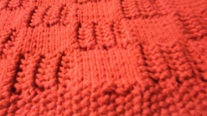 May include: Close-up of a red knitted fabric with a textured pattern. The fabric is soft and fluffy, and the pattern is created by the way the yarn is woven together.
