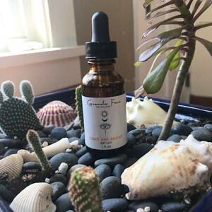 Puede incluir: Una botella de vidrio marr&oacute;n de suero Light and Bright de Granola Face Skincare con un cuentagotas negro. La botella est&aacute; colocada en una jardinera azul con cactus, conchas marinas y piedras grises. La etiqueta tiene un dise&ntilde;o de luna y ojo.