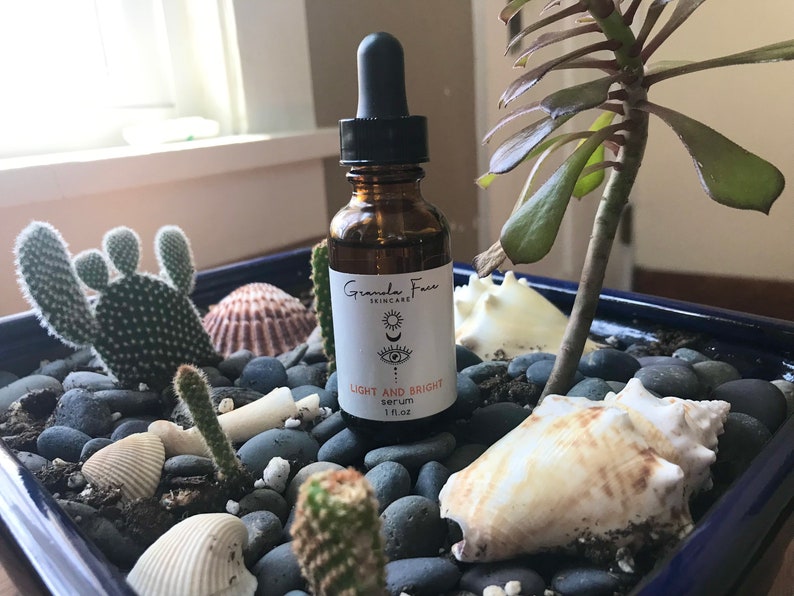 Puede incluir: Una botella de vidrio marr&oacute;n de suero Light and Bright de Granola Face Skincare con un cuentagotas negro. La botella est&aacute; colocada en una jardinera azul con cactus, conchas marinas y piedras grises. La etiqueta tiene un dise&ntilde;o de luna y ojo.