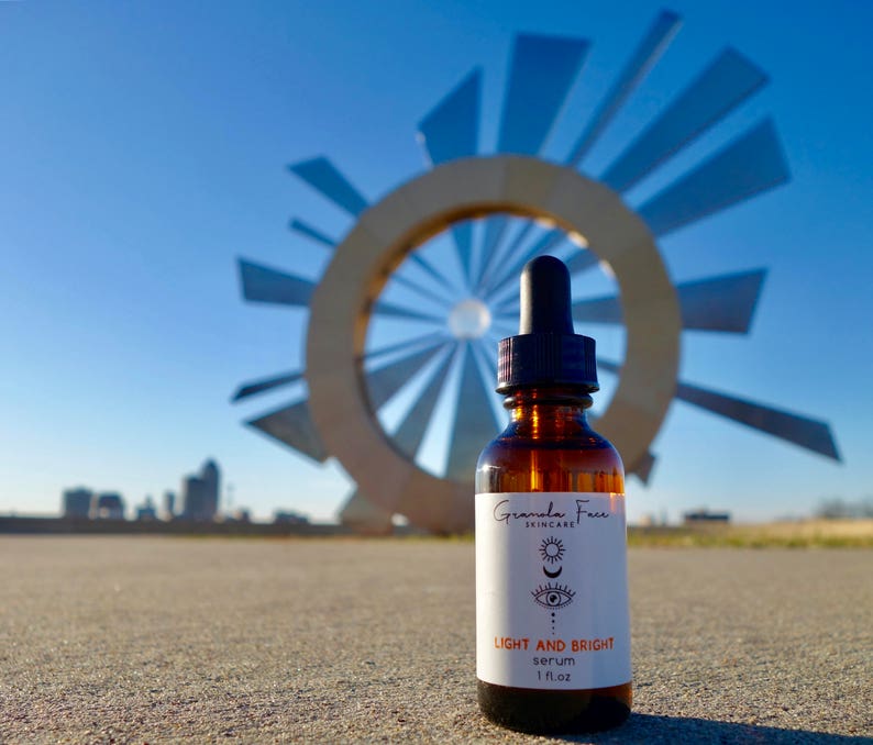 Puede incluir: Una botella de vidrio marr&oacute;n de suero Granola Face Skincare con un cuentagotas, etiquetada como "LIGHT AND BRIGHT serum 1 fl.oz.". La botella se encuentra frente a una gran escultura de metal y un cielo azul claro.