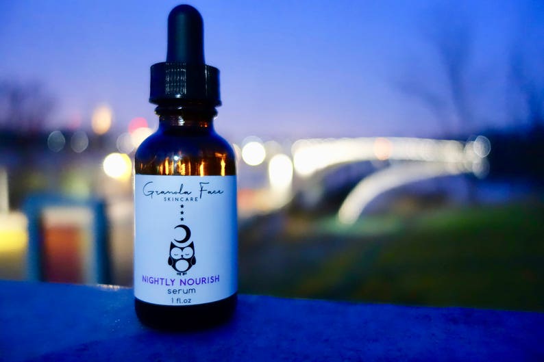 May include: A brown glass serum bottle with a black dropper. The label reads "Granola Face Skincare" and "Nightly Nourish serum 1 fl oz." An owl and moon graphic is on the label. The background is blurred with city lights.