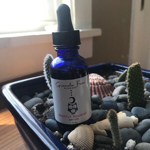 May include: A blue glass serum bottle with a black dropper, labeled "Nightly Nourish serum 1 fl oz." The bottle is in a blue square planter with rocks, seashells, and cacti. The brand name is "Granda Face Skincare."