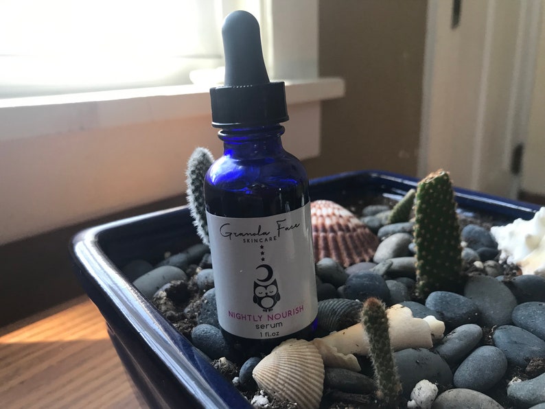 May include: A blue glass serum bottle with a black dropper, labeled "Nightly Nourish serum 1 fl oz." The bottle is in a blue square planter with rocks, seashells, and cacti. The brand name is "Granda Face Skincare."