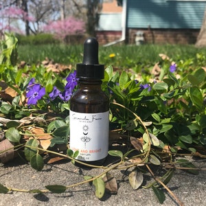 Puede incluir: Un frasco de suero de vidrio marr&oacute;n con una tapa cuentagotas negra, etiquetado como "Granola Face Skincare". La etiqueta presenta un dise&ntilde;o de ojo y luna, con las palabras "LIGHT AND BRIGHT serum" y "1 fl oz". La botella est&aacute; rodeada de follaje verde y flores moradas.