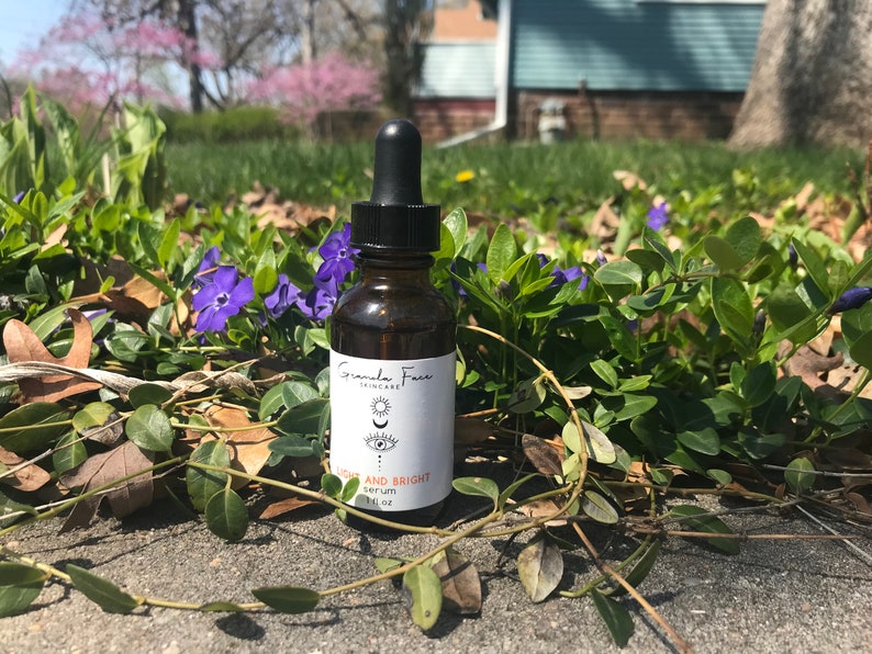 Puede incluir: Un frasco de suero de vidrio marr&oacute;n con una tapa cuentagotas negra, etiquetado como "Granola Face Skincare". La etiqueta presenta un dise&ntilde;o de ojo y luna, con las palabras "LIGHT AND BRIGHT serum" y "1 fl oz". La botella est&aacute; rodeada de follaje verde y flores moradas.