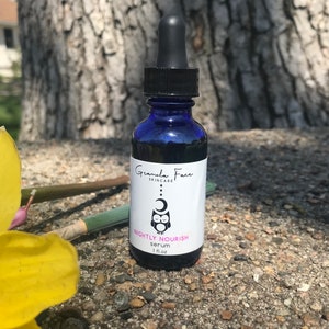 May include: A blue glass serum bottle with a black dropper. The label reads "Granola Face Skincare" and "Nightly Nourish serum 1 fl oz". An owl and moon graphic is featured on the label. The bottle is set against a natural background.