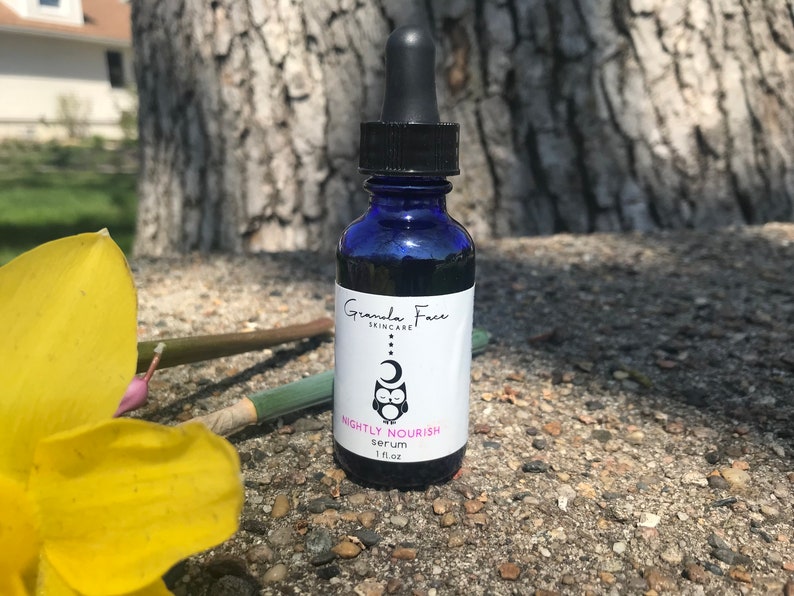 May include: A blue glass serum bottle with a black dropper. The label reads "Granola Face Skincare" and "Nightly Nourish serum 1 fl oz". An owl and moon graphic is featured on the label. The bottle is set against a natural background.