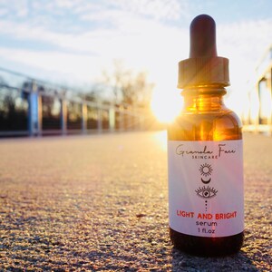Puede incluir: Botella de vidrio marr&oacute;n de s&eacute;rum "Light and Bright" de "Granola Face Skincare" con cuentagotas. La etiqueta presenta un sol, una luna y un ojo. La botella es de 30 ml. El fondo es una escena exterior soleada.