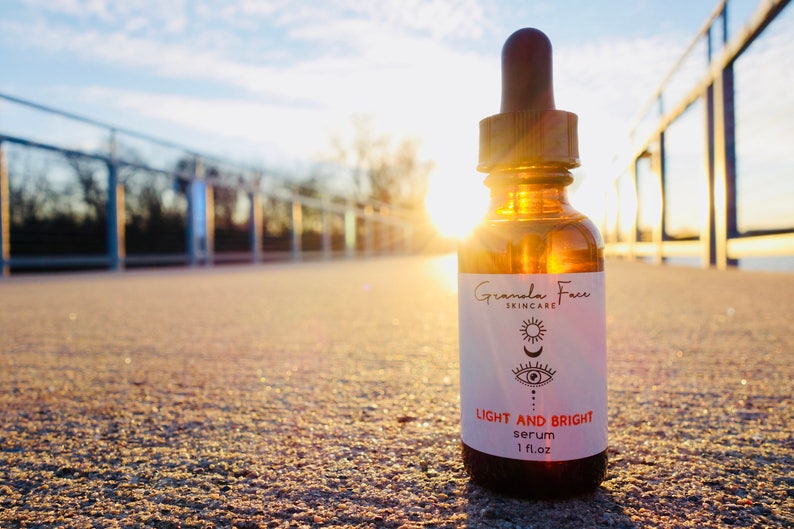 Puede incluir: Botella de vidrio marr&oacute;n de s&eacute;rum "Light and Bright" de "Granola Face Skincare" con cuentagotas. La etiqueta presenta un sol, una luna y un ojo. La botella es de 30 ml. El fondo es una escena exterior soleada.