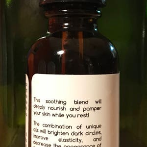 May include: A dark amber glass bottle with a black dropper top. The bottle has a white label with text describing a soothing blend of oils to nourish and pamper skin, reduce wrinkles, and improve elasticity.