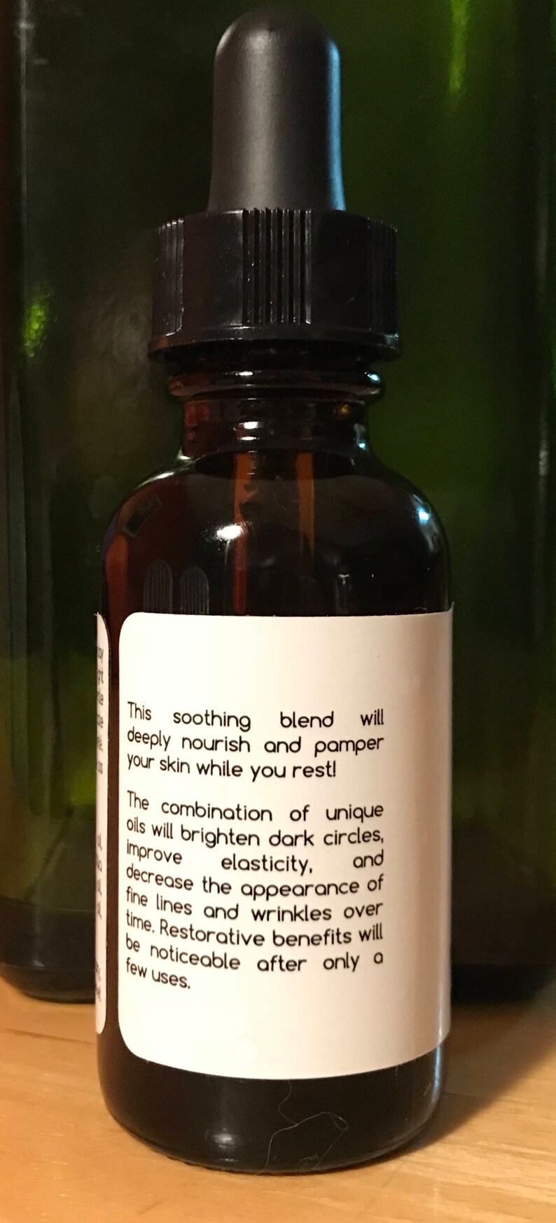 May include: A dark amber glass bottle with a black dropper top. The bottle has a white label with text describing a soothing blend of oils to nourish and pamper skin, reduce wrinkles, and improve elasticity.