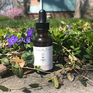 Puede incluir: Una botella de suero de vidrio marr&oacute;n con un cuentagotas negro, sobre un fondo de vegetaci&oacute;n y flores moradas. La etiqueta dice "Granola Face Skincare" con las palabras "Light and Bright Serum".