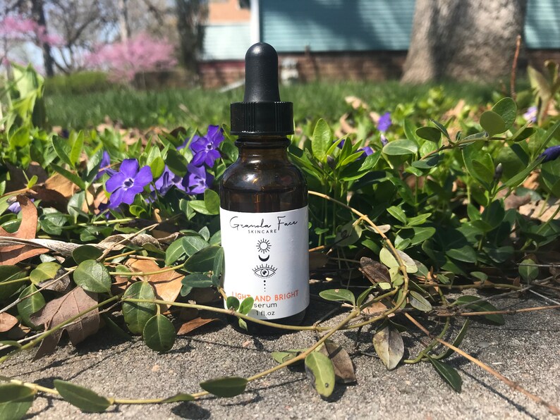 Puede incluir: Una botella de suero de vidrio marr&oacute;n con un cuentagotas negro, sobre un fondo de vegetaci&oacute;n y flores moradas. La etiqueta dice "Granola Face Skincare" con las palabras "Light and Bright Serum".