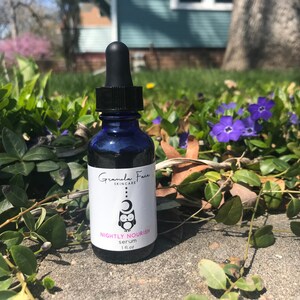 May include: A dark blue glass bottle with a black dropper, labeled "Nightly Nourish serum" with a logo of an owl and moon. The bottle contains 1 fl oz of serum. The bottle is set against a backdrop of green foliage and purple flowers.