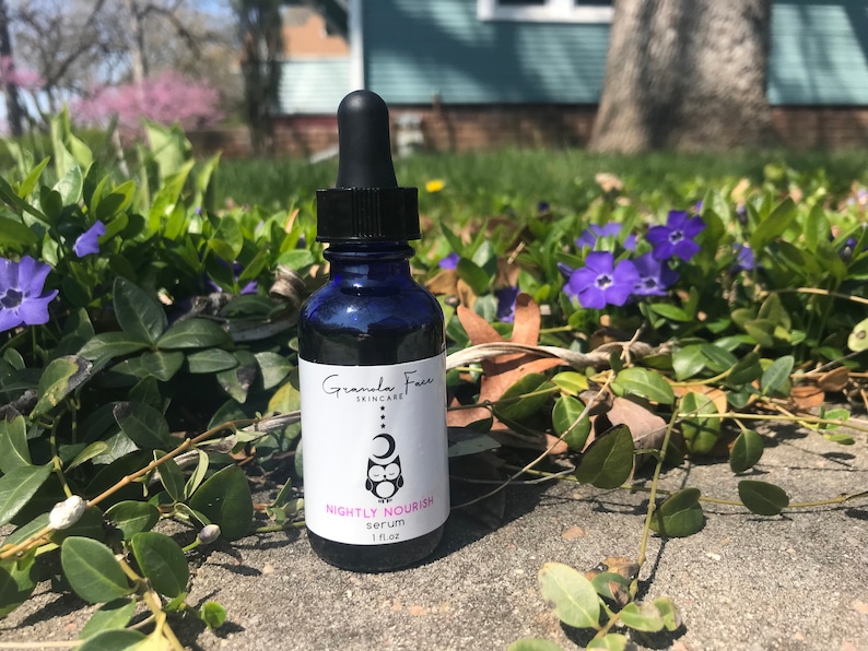 May include: A dark blue glass bottle with a black dropper, labeled "Nightly Nourish serum" with a logo of an owl and moon. The bottle contains 1 fl oz of serum. The bottle is set against a backdrop of green foliage and purple flowers.