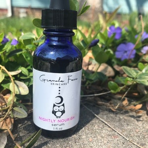 May include: A dark blue glass bottle of Granola Face Skincare Nightly Nourish serum. The label features an owl, moon, and stars. The bottle has a black dropper and is set against a background of green foliage and purple flowers. The bottle contains 1 fl oz of serum.