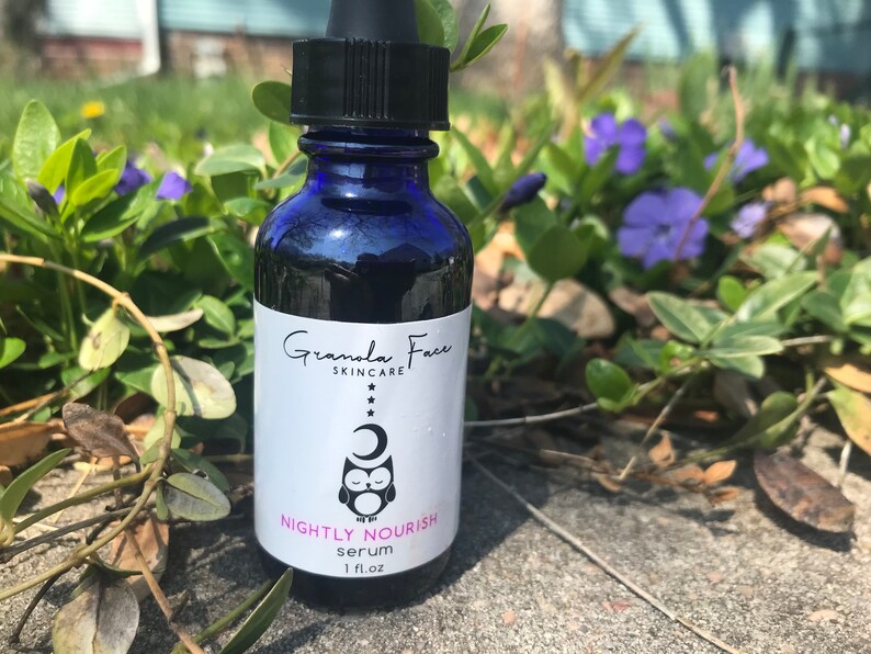 May include: A dark blue glass bottle of Granola Face Skincare Nightly Nourish serum. The label features an owl, moon, and stars. The bottle has a black dropper and is set against a background of green foliage and purple flowers. The bottle contains 1 fl oz of serum.
