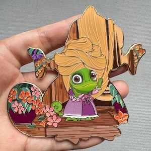 May include: A gold-tone enamel pin featuring a green chameleon wearing a purple dress and a blonde wig. The chameleon is standing on a wooden platform with flowers and greenery surrounding it.