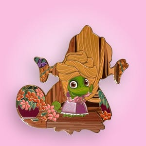 May include: A metal enamel pin featuring a green frog character with large eyes, wearing a pink and purple dress and a large tan head wrap. The frog is positioned in front of a wooden door frame with a potted plant containing orange flowers.