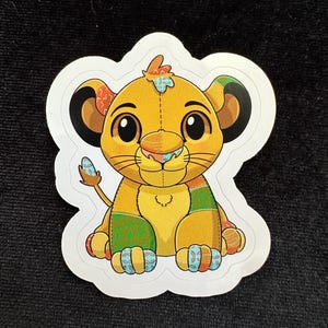 Puede incluir: Una pegatina de un lindo cachorro de león de estilo de dibujos animados con un diseño de patchwork. El león está sentado con las patas cruzadas y tiene una expresión amigable. La pegatina es amarilla, verde, azul y naranja.
