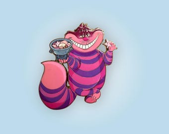 May include: A colorful enamel pin featuring the Cheshire Cat from Alice in Wonderland. The pin depicts the cat with a wide grin, holding a bowl of treats and a star-shaped object. The cat has pink and purple stripes and a long tail.