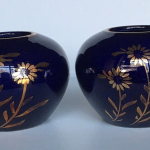 May include: Two small, cobalt blue ceramic candle holders with gold floral designs. The holders are round and have a slightly flared lip.