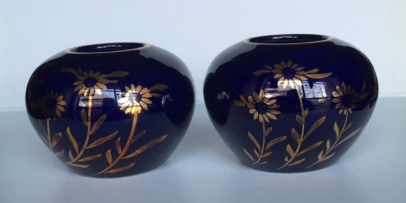 May include: Two small, cobalt blue ceramic candle holders with gold floral designs. The holders are round and have a slightly flared lip.