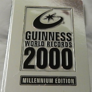 May include: A silver-colored book titled "Guinness World Records 2000 Millennium Edition." The cover features a black logo with a crescent and star, and the title in bold, black lettering. The book is rectangular and appears to be in good condition.