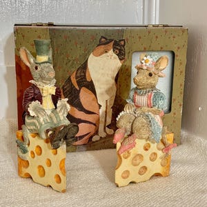 May include: A decorative box with a painted cat and two rabbit figurines sitting on cheese wedges. The rabbits are dressed in Victorian-era clothing. The box has a green floral pattern.