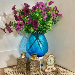 May include: A vibrant still life featuring a turquoise blue glass vase filled with purple flowers and green foliage. Two ornate perfume bottles with gold accents and a small clock with floral details sit on a lace doily.