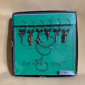 May include: A set of six golf-themed wine glass charms in a black box with a green background. The charms feature a golf bag, golf clubs, a golf ball, a martini glass, a golf tee, and a golfer. The box has the text "that wine is mine!™" and "fore!" printed on it.