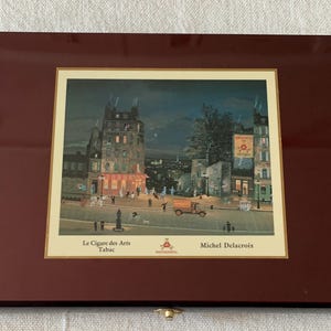 May include: A wooden box with a hinged lid, featuring a framed print of a Parisian street scene at night. The print depicts a building with a sign that reads "Le Cigare des Arts Tabac" and is signed "Michel Delacroix".