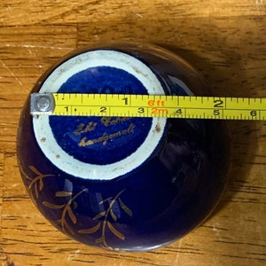 May include: A small, cobalt blue ceramic bowl with gold leaf detailing. The bowl is approximately 3 inches in diameter and has a white interior. The bottom of the bowl is marked with the text "Lili Cohrs Handgemalt".