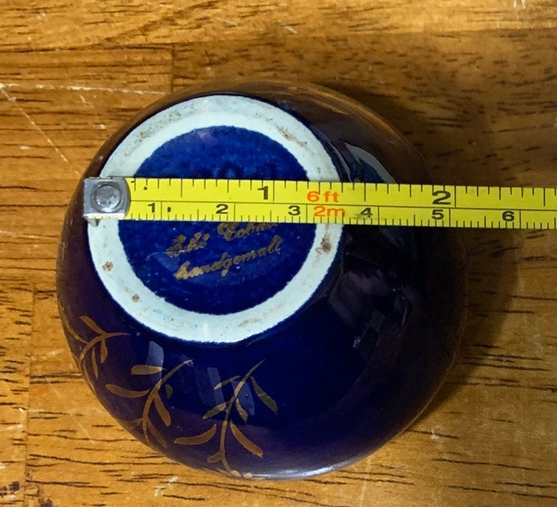 May include: A small, cobalt blue ceramic bowl with gold leaf detailing. The bowl is approximately 3 inches in diameter and has a white interior. The bottom of the bowl is marked with the text "Lili Cohrs Handgemalt".
