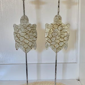 May include: Two decorative metal candle holders. Each has a tall, slender stem with a turtle-shaped cutout and a small, round candle platform. The turtles are painted in a distressed white and brown finish, and the bases are green.
