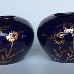 May include: Two small, cobalt blue ceramic candle holders with gold floral designs. The holders are round and have a slightly flared lip.