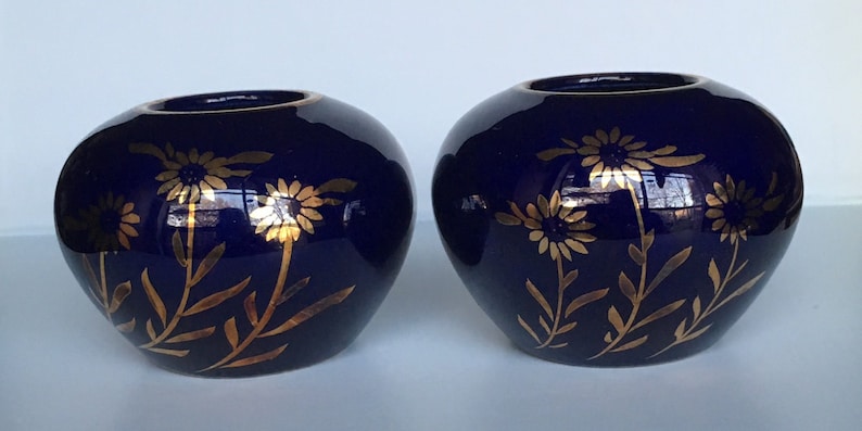 May include: Two small, cobalt blue ceramic candle holders with gold floral designs. The holders are round and have a slightly flared lip.